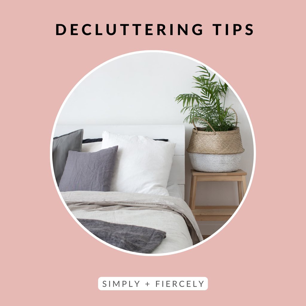 A round image of a bed with white sheets and a grey blanket, fluffy pillows, and a side table with a plant on it on a pink background with the words Decluttering Tips across the top.