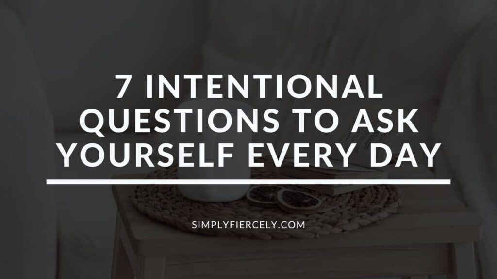 "7 Intentional Questions To Ask Yourself Every Day" in white letters on a translucent black overlay on top of an image of a table with a coffee cup, orange slice, a book, and a wicker mat on top.