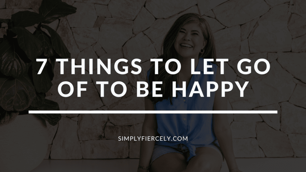 "7 Things To Let Go Of To Be Happy" in white letters on a translucent black overlay on top of an image of a smiling woman wearing a blue sleeveless top and denim shorts sitting in front of a stone wall.