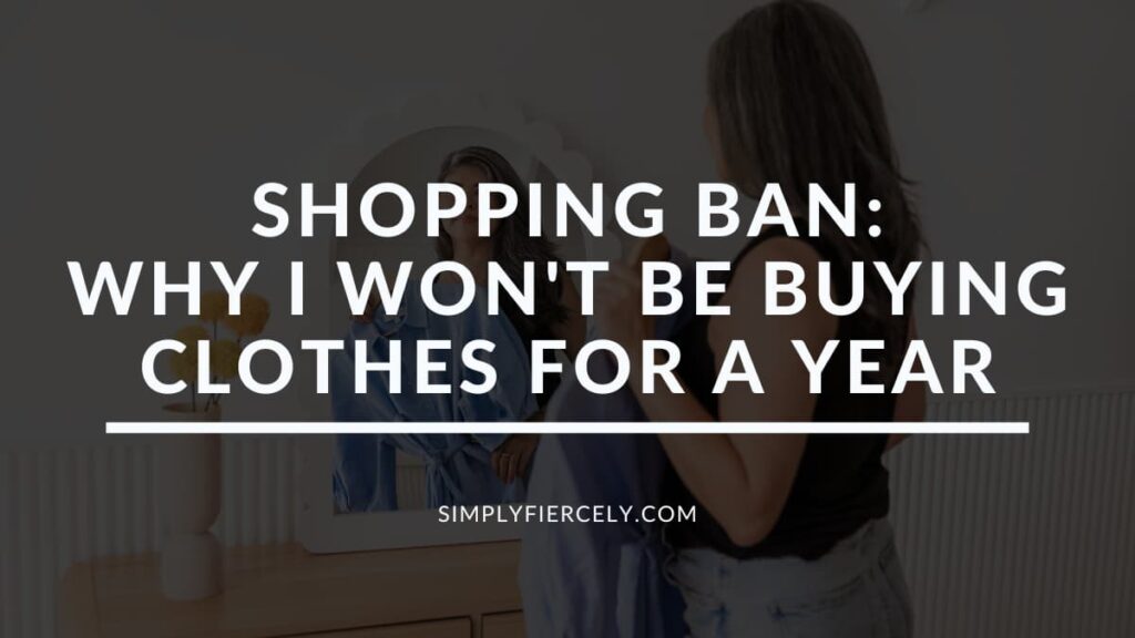"Shopping Ban: Why I Won't Be Buying Clothes For A Year" in white letters on a translucent black overlay on top of an image of a woman wearing a sleeveless black top and denim shorts holding a blue dress up to her while looking it the mirror.