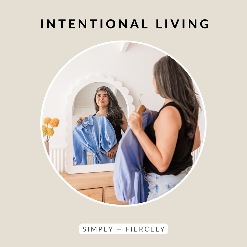 A round image of a smiling woman wearing a sleeveless black top and denim shorts holding a blue dress up to her while looking it the mirror on a beige background with the words Intentional Living across the top.