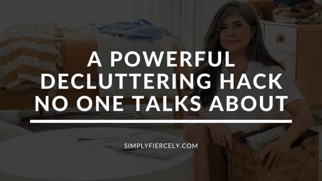 "A Powerful Decluttering Hack No One Talks About" in white lettering on a translucent black overlay on an image of a smiling woman holding a woven basket with books inside sitting on the floor in a room full of clutter.