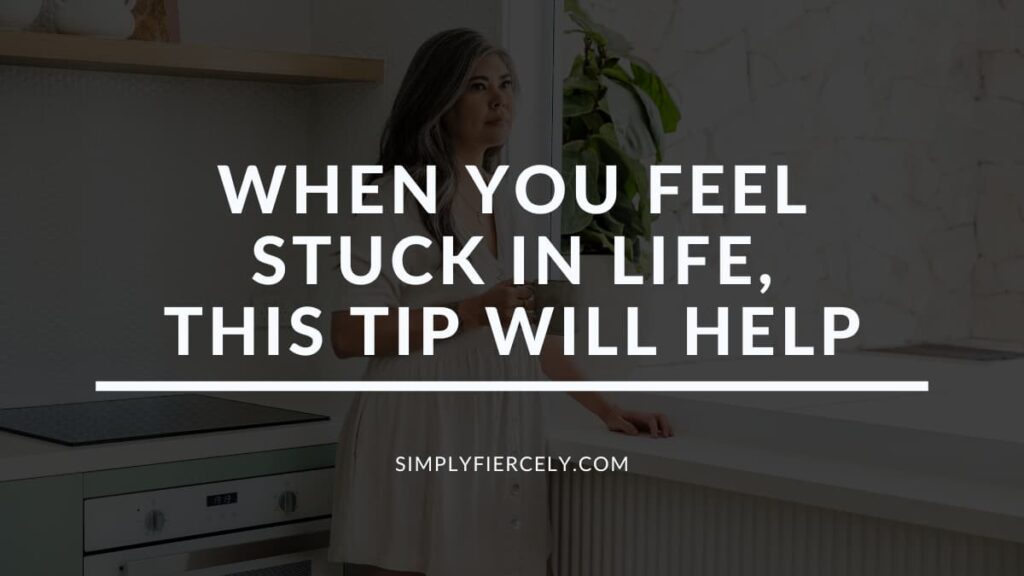 "When You Feel Stuck In Life, This Tip Will Help You Move Forward" in white letters on a translucent black overlay on top of an image of a woman wearing a loose white dress holding a coffee cup in a kitchen looking out a window.