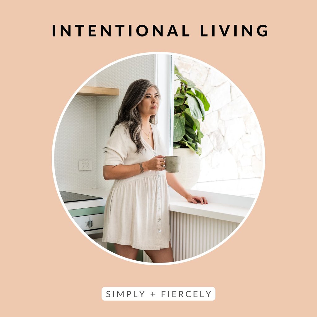 A round image of a woman wearing a loose white dress holding a coffee cup in a kitchen looking out a window on an orange background with the words Intentional Living across the top