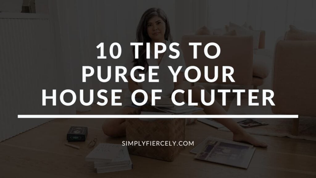 "10 Tips to Purge Your House of Clutter" in white letters on a translucent black overlay on top of an image of a woman wearing shorts and a white button up top smiling at the camera while sitting on the floor decluttering a basket.